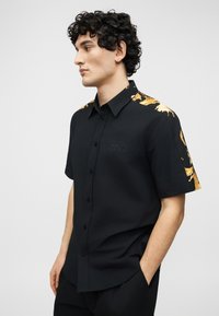 Άνδρας με σγουρά μαλλιά που φοράει μαύρο πουκάμισο με κοντά μανίκια και χρυσό μπαρόκ σχέδιο στους ώμους και τα μανίκια, στέκεται με τα χέρια στις τσέπες.