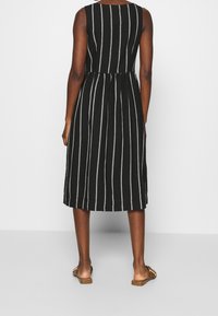 Robe sans manches à rayures noires et blanches avec une taille cintrée, une longueur jusqu'aux genoux et un tissu léger. Portée avec des sandales plates.