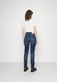 Person med lockigt hår som bär en vit t-shirt, mörkblå skinny jeans med en logopatch och svarta skor, står vänd mot en plain vit vägg.
