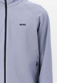 Lys grå jakke med glidelås laget av glatt stoff. Har svart glidelås og logo på brystet, med lommer foran på hver side.