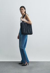 Mustat tyynyttävät kangaskassit, joissa on jämäkkä muotoilu, pehmeä, vanutettu pinta ja leveät hihnat. Mallilla on päällään vaaleansiniset farkut ja korkokengät.