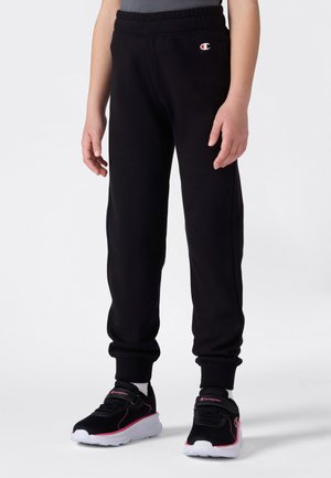 Barn som har på seg svarte Champion joggebukser og svarte sneakers med hvite såler og rosa detaljer på hvit bakgrunn.
