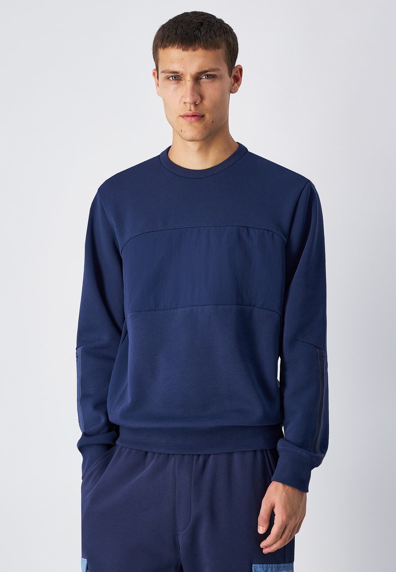Felpa blu navy con un pannello testurizzato sul petto, maniche lunghe, scollatura a girocollo e polsini a costine. Abbinata a pantaloni blu navy coordinati.