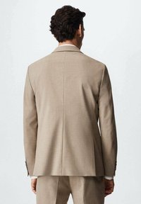 Giacca da completo beige con una texture liscia, colletto rigido, spacco singolo sul retro e dettagli di polsini abbottonati.