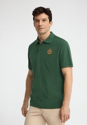 Homme portant un polo vert foncé avec un écusson brodé sur la poitrine gauche, associé à un pantalon beige, debout sur un fond blanc.