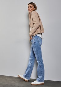 Chaqueta ligera beige con puños elásticos, camiseta blanca, jeans acampanados de color azul claro y zapatillas blancas con suelas marrones sobre un fondo gris.