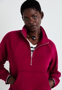 Sweat à zip fuchsia avec un col haut, des poignets côtelés et une poche frontale. porté par-dessus une chemise rayée noir et blanc.