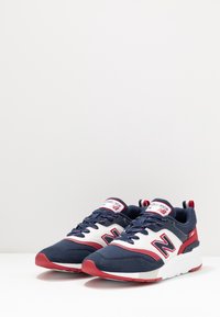 Chaussures de sport bleues et blanches avec des accents rouges, dotées d'une tige texturée, d'une languette rembourrée et d'un marquage bien visible sur le côté.