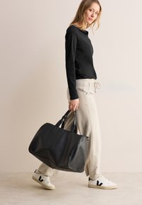 Zwarte leren sporttas met dubbele handvatten, gladde textuur en ronde vorm. Model draagt een zwart shirt met lange mouwen en beige broek.
