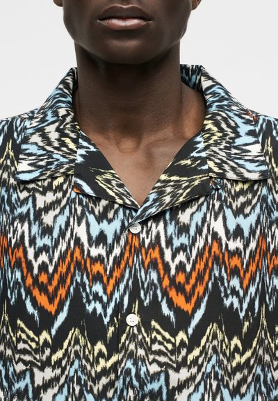 Κοντινό πλάνο ενός ατόμου που φοράει πουκάμισο με κουμπιά με έντονο ζιγκ ζαγκ μοτίβο σε μπλε, πορτοκαλί, μαύρο, κρεμ και λευκό χρώμα.