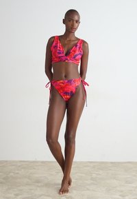 Dvoslojni bikini set v živahnih roza, rdečih in vijoličnih cvetličnih vzorcih. Zgornji del ima V-izrez, spodnji del pa ima zavezujoče detajle. Gladka tekstura.