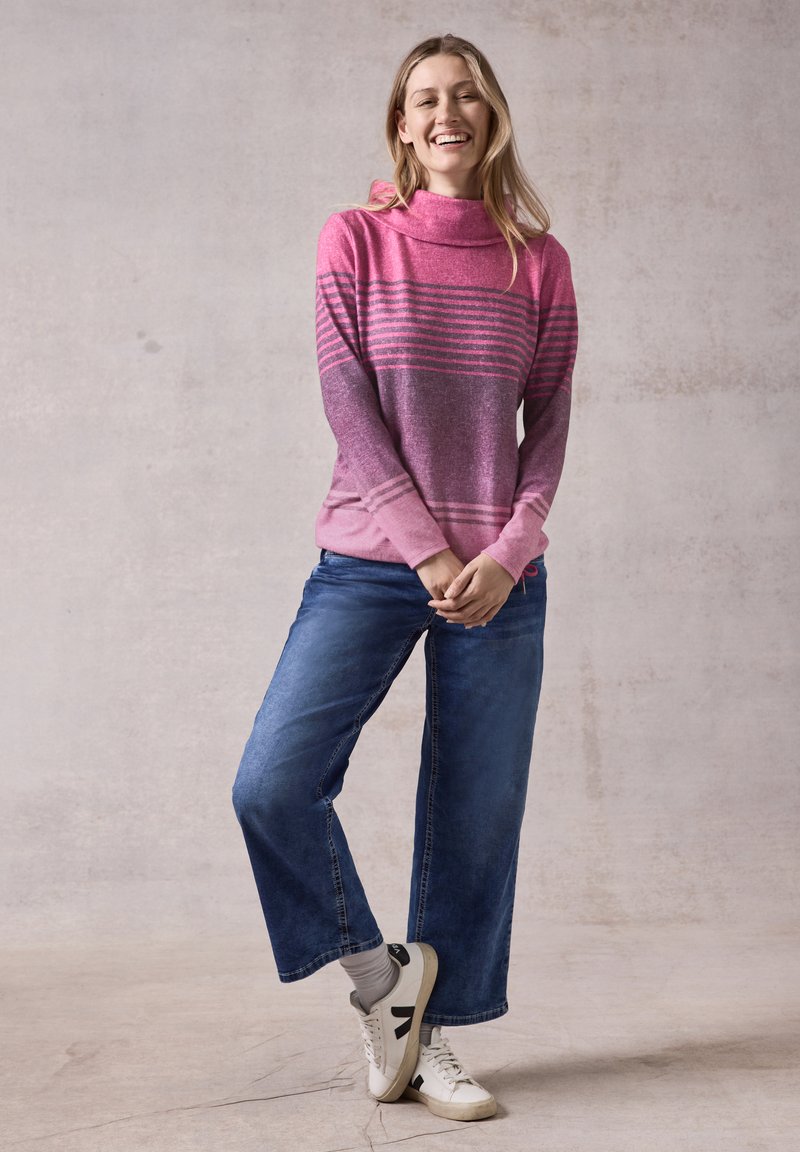 Rosa und lila gestreifter Pullover mit einem Schalkragen, kombiniert mit weiten blauen Jeans und weißen Sneakern. Weicher Stoff und lässiges Design.