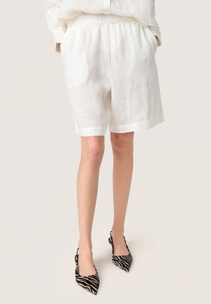 Személy laza fehér len rövidnadrágot és zebramintás hegyes orrú slingback magas sarkú cipőt visel, zsebébe tett kézzel áll.
