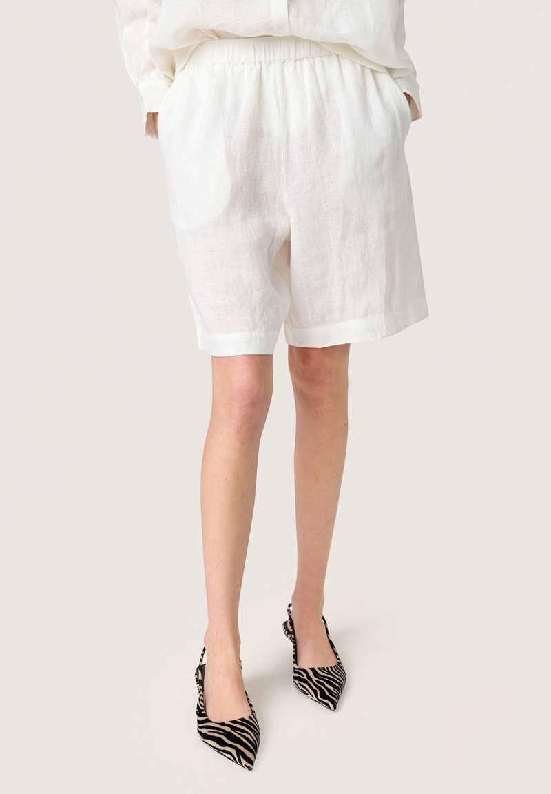 Personne portant un short ample en lin blanc et des escarpins slingback à bout pointu imprimés zèbre, debout les mains dans les poches.