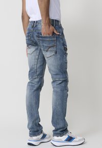 Unos vaqueros de mezclilla azul con un diseño ajustado, cinco bolsillos y detalles de costura visibles, combinados con zapatillas blancas y una camiseta blanca lisa.