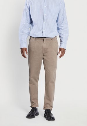 Mann trägt beige plissierte Hose, schwarze Lederschuhe und ein hellblau gestreiftes Langarmhemd vor einfachem Hintergrund.