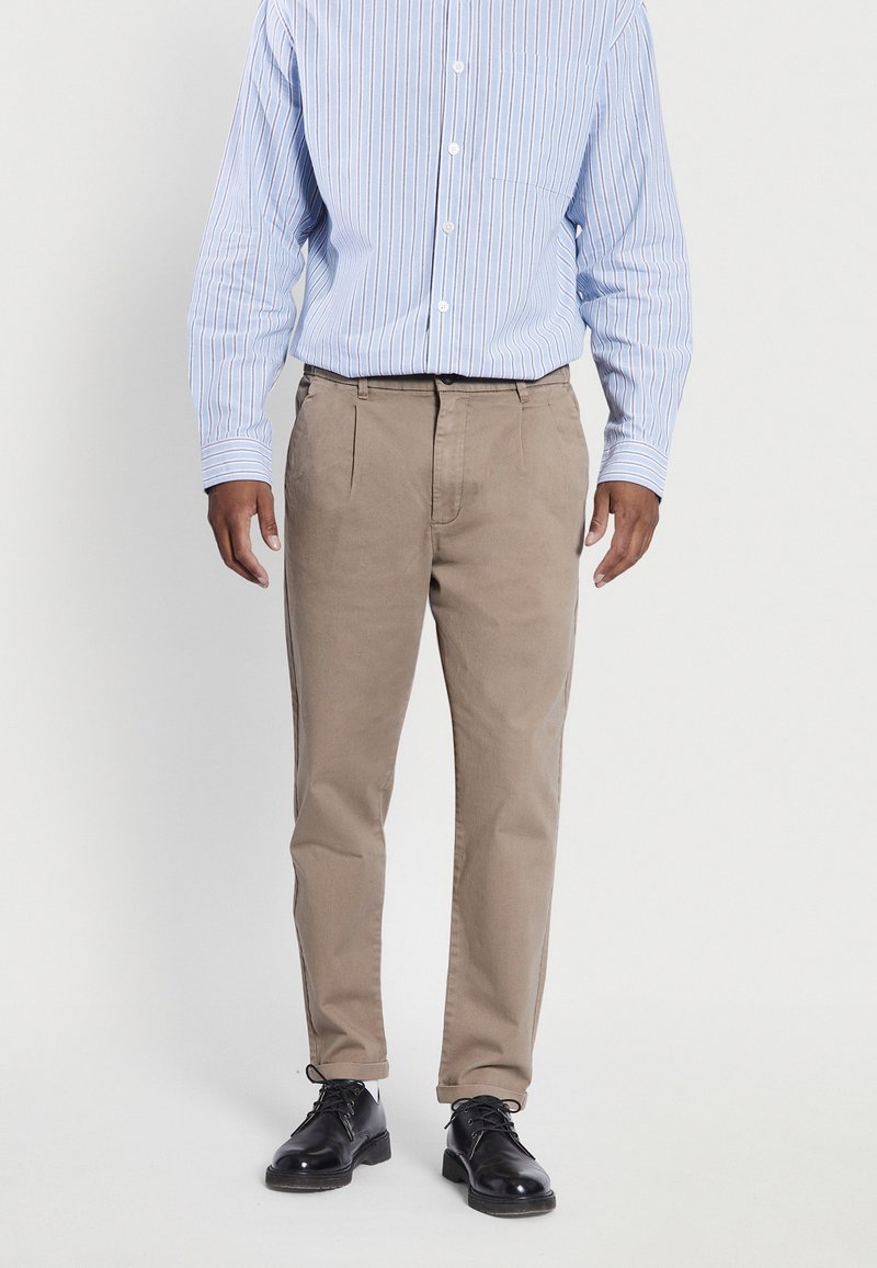 Homme portant un pantalon beige plissé, des chaussures de ville noires et une chemise à manches longues rayée bleu clair sur un fond uni.