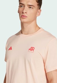 Světlá růžová sportovní tričko vyrobené z hladké tkaniny, s červeným logem Adidas a výrazným emblémem "AR" na hrudi a rukávu.