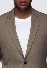 Brązowa marynarka w jodełkę z jednym guzikiem, klapami z wcięciem i kieszenią na piersi, noszona na białej koszulce. Widoczna tekstura tkaniny.