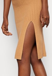 Virkat klänning i en varm beige färg med ribbad textur, med en sidoklyfta som går till låret. Närbild av hand- och ben-detalj.