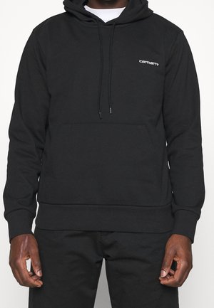 Sweat à capuche noir Carhartt avec poche avant et cordons, porté par une personne à la peau foncée et portant un pantalon noir.