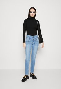 Gaiši zili augsto jostasvietu džinsi ar pogu priekšā, kombinēti ar melnu augstu apkakli un masīviem melniem loaferi ar zelta akcentiem.