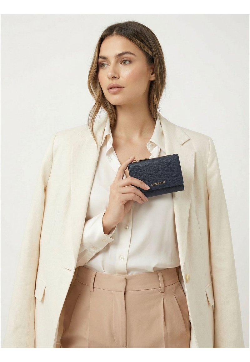 Mujer con blazer y pantalones beige sosteniendo una pequeña cartera azul marino con letras doradas "LAZAROTTI" cerca de su pecho.
