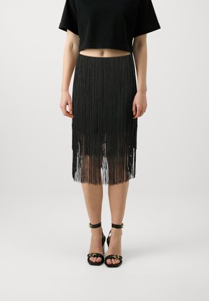 Femme portant une jupe noire à franges, un top court noir et des sandales noires à bride cheville, debout devant un fond uni.