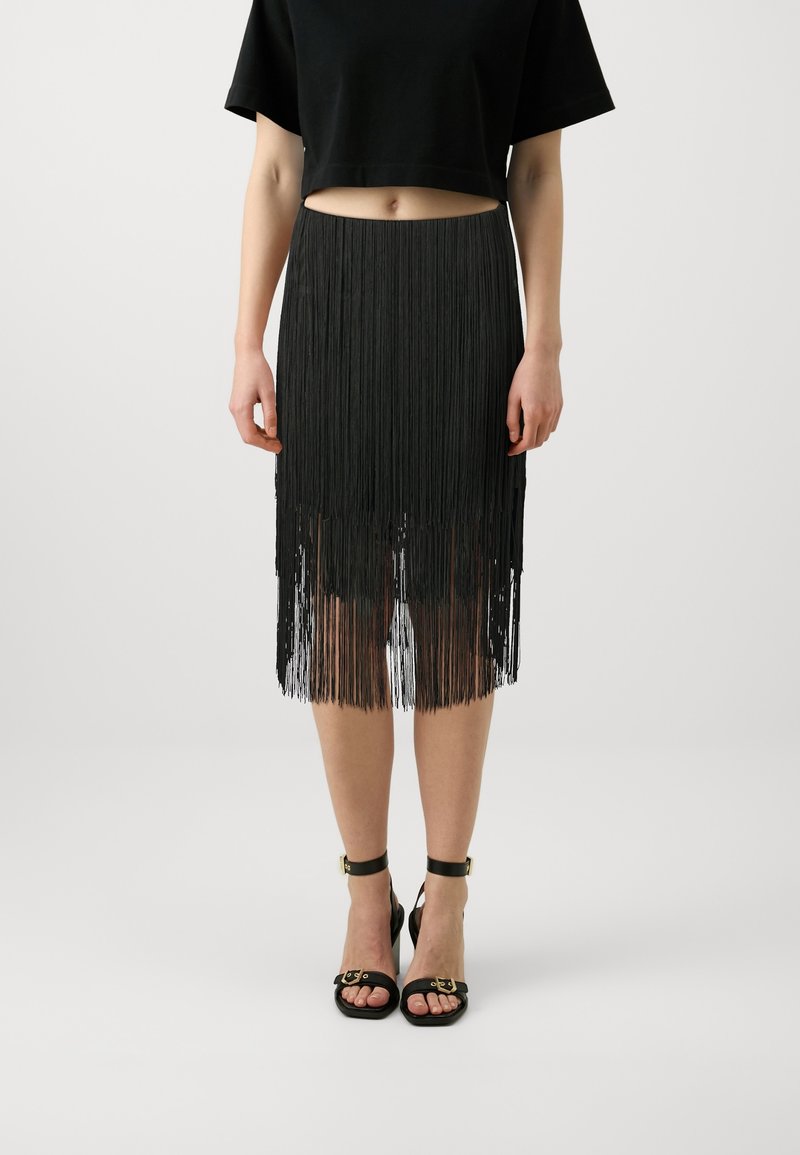 Femme portant une jupe noire à franges, un top court noir et des sandales noires à bride cheville, debout devant un fond uni.