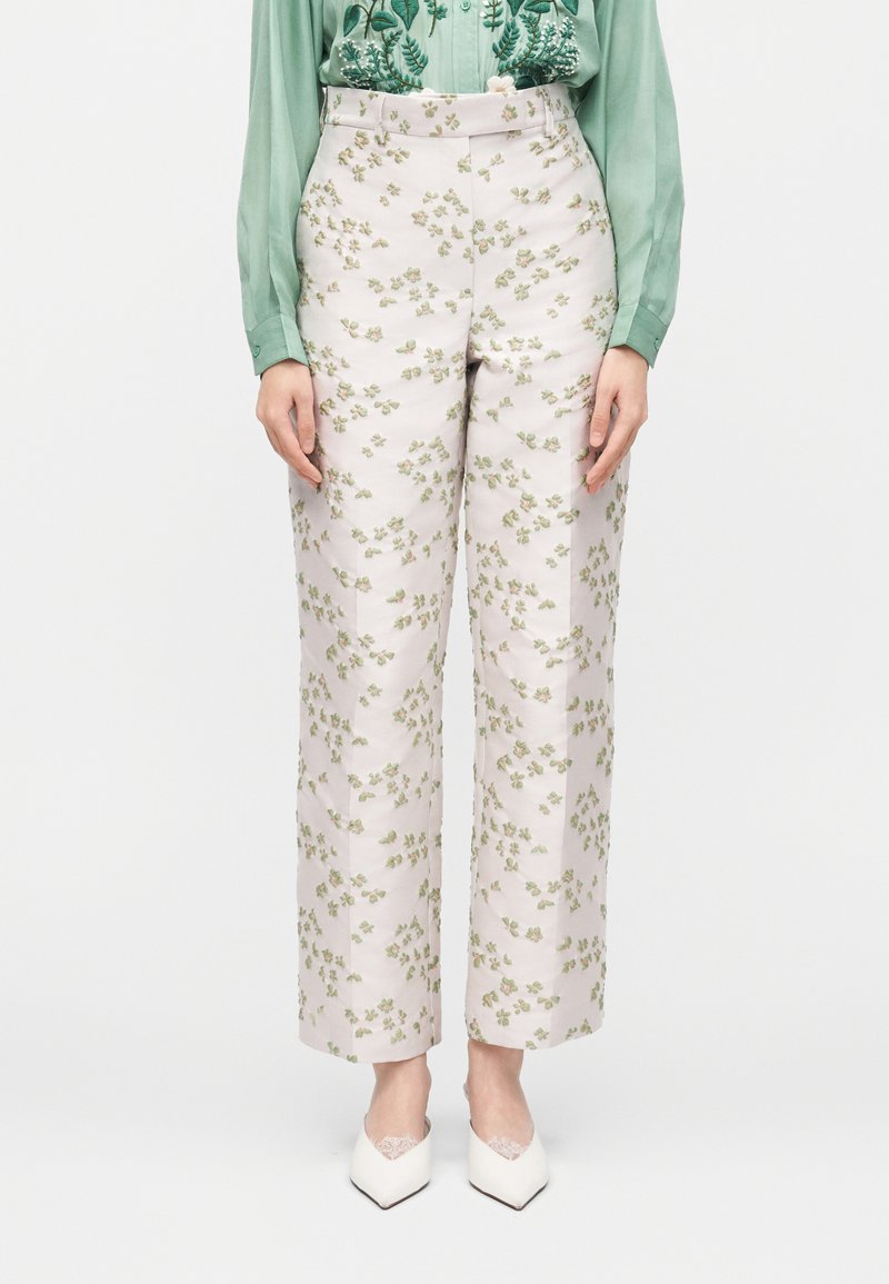 Γυναίκα με ψηλόμεσο άσπρο παντελόνι με πράσινο λουλουδάτο σχέδιο, συνδυασμένο με πράσινο πουκάμισο με κουμπιά και λευκές μυτερές γόβες.