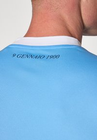 Vaaleansininen urheilupaita, jossa on valkoinen kaulus. Teksti "9 GENNAIO 1900" painettuna mustana selässä, kauluksen alapuolella. Sujuva kangastuntuma.