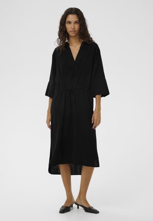 Kobieta o kręconych włosach, ubrana w czarną sukienkę midi z rękawami 3/4, dekoltem w serek oraz czarne mule na niskim obcasie, stojąca na jasnym, jednolitym tle.