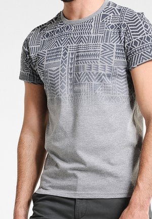 Mężczyzna w szarej koszulce z krótkim rękawem z ciemnymi geometrycznymi wzorami na górnej części klatki piersiowej i na rękawach.