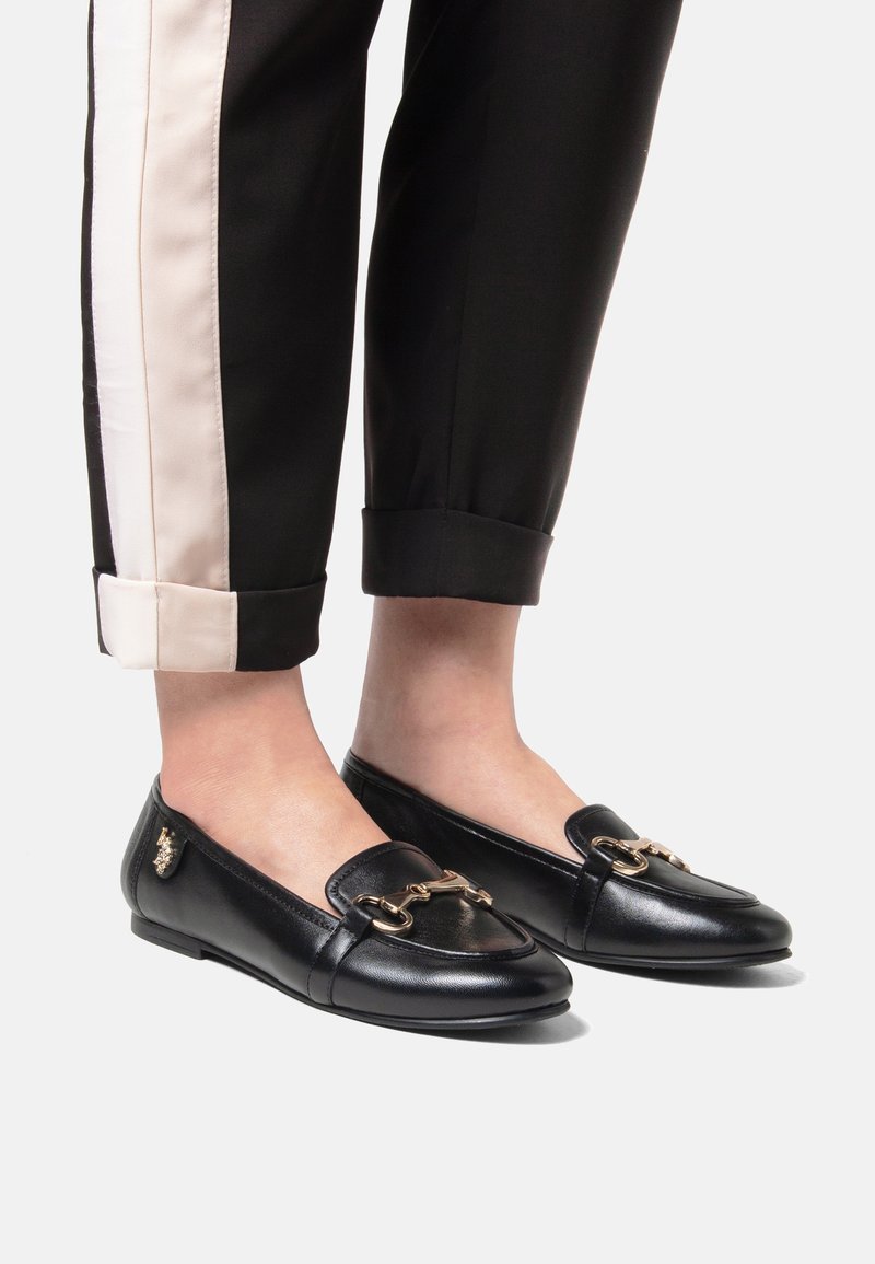 Svarta läderloafers med låg profil, utrustade med en guldtonad hästbitdetalj och slät yta, matchade med svarta byxor med en ljus rand.