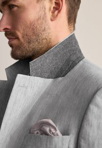 Helle graue Jacke mit einem kontrastierenden dunkelgrauen Wollkragen. Vorgestelltes Taschentuch in helllila. Strukturierten Stoff mit klaren Linien.