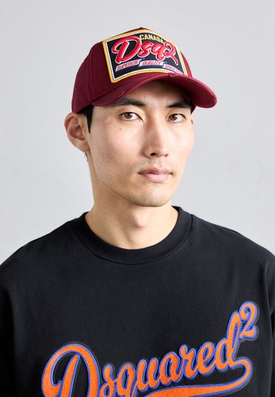 Young man wearing a burgundy cap with a Canada Dsquared2 logo and a black sweatshirt with bold orange and blue Dsquared2 text.