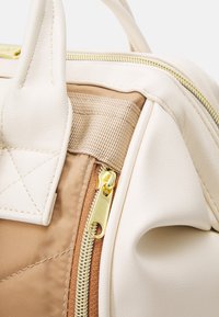 Gros plan sur un sac texturé beige et marron clair montrant une fermeture éclair dorée, une poignée cousue et un détail de boucle en tissu.