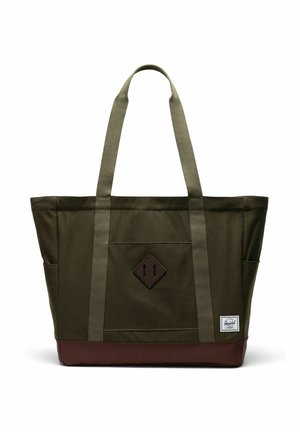 Sac fourre-tout en toile vert olive avec base marron, deux longues anses, poche avant, et petite étiquette de marque blanche rectangulaire en bas à droite.
