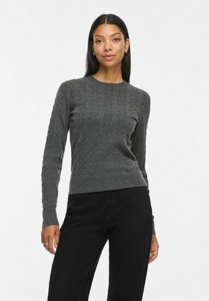 Woman with long dark hair wears a fitted dark gray cable knit sweater and black pants standing against a plain white background.