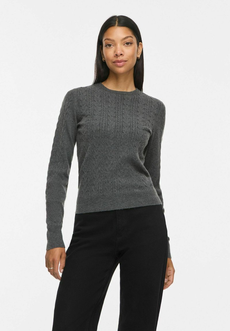 Femme aux longs cheveux foncés portant un pull en maille torsadée gris foncé ajusté et un pantalon noir, debout devant un fond blanc uni.