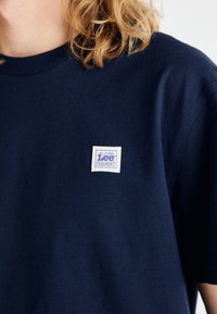 T-shirt en coton bleu marine avec un col rond et des manches courtes. Il présente une étiquette en tissu carrée blanche avec du texte bleu sur la poitrine gauche.