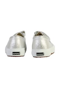 Zapatillas de lona plateadas brillantes con suela de goma blanca, que cuentan con un diseño de cordones y el logotipo de la marca en el talón. Se muestra la vista trasera de los zapatos.