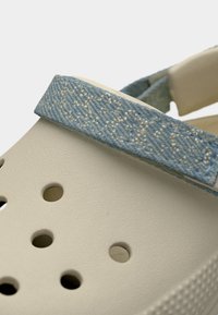 Sandale beige avec des trous de ventilation et une sangle en tissu bleu texturé ajustable sur le dessus.