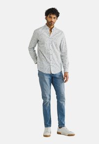 Witte overhemd met blauwe en groene stippen, gecombineerd met lichtblauwe jeans en witte sneakers met bruine zolen. Casual pasvorm.