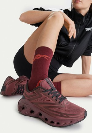 Persona atlética con ropa negra sentada con una pierna doblada, usando zapatillas Reebok color burdeos y calcetines a juego sobre fondo blanco.