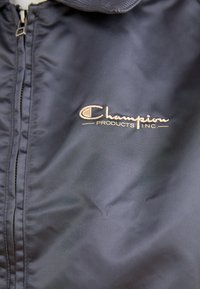 Črna jakna na zadrgo iz gladkega materiala; ima vezeno zlato "Champion" logo na levi strani.
