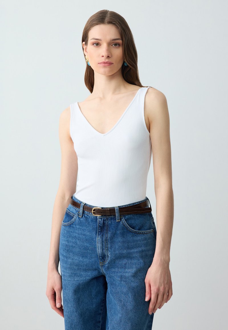 Femme aux longs cheveux bruns portant un top blanc sans manches à col en V, un jean bleu taille haute et une ceinture marron, debout devant un fond uni.