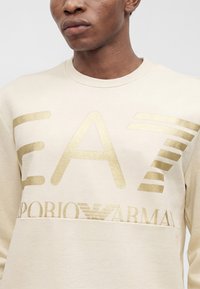Κοντινό πλάνο ενός ατόμου που φοράει κρεμ φούτερ με μεγάλο χρυσό λογότυπο EA7 Emporio Armani στο μπροστινό μέρος.