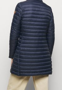 Marine pufferjack met horizontaal gestikte patroonen, lange mouwen, en een licht uitlopende zoom. Heeft een embleem op de linkerschouder.