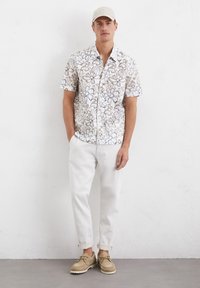 Korte mouwen bloemenoverhemd in wit en bruin, gecombineerd met opgerolde lichte broeken en beige schoenen. Het overhemd heeft een button-down kraag.